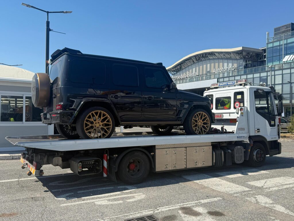 Sul pianale del carro attrezzi è caricato un grande SUV di lusso nero lucido. Il SUV presenta vetri oscurati, vistosi cerchi in lega multirazze di colore oro/bronzo e una copertura della ruota di scorta posteriore dello stesso colore metallizzato.