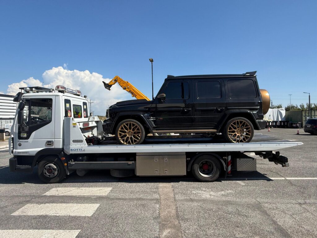 Sul pianale del carro attrezzi è caricato un grande SUV di lusso nero lucido. Il SUV presenta vetri oscurati, vistosi cerchi in lega multirazze di colore oro/bronzo e una copertura della ruota di scorta posteriore dello stesso colore metallizzato.